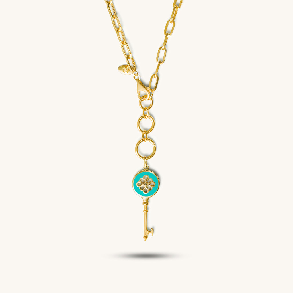 A 18k gold necklace with a golden key pendant featuring a blue enamel center with a floral motif, attached to a rectangular chain.